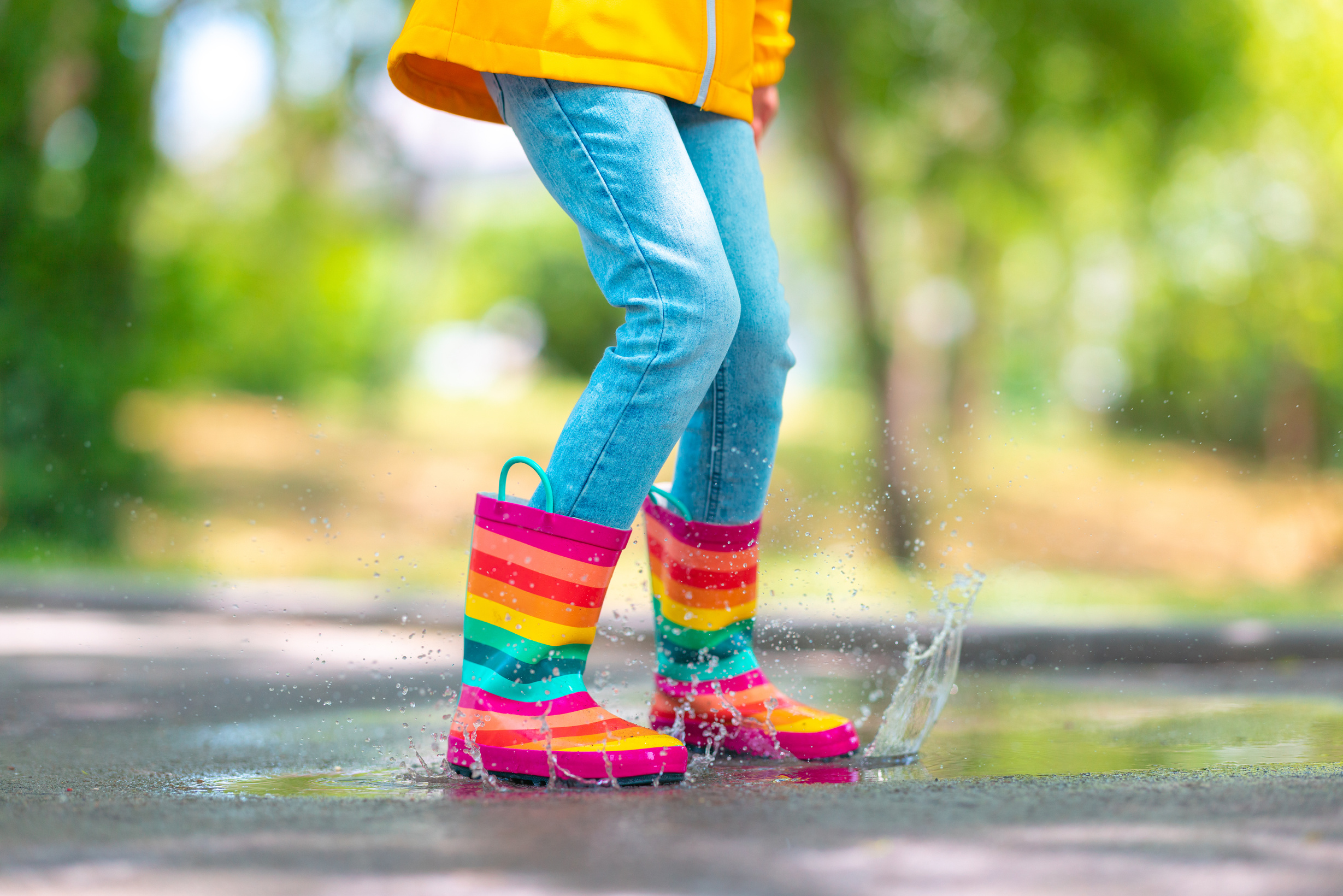 Feet of child in colorful  rubber rain boots jumping over rainy puddle in a park